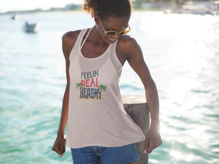 Lifestyle mockup of the funny, travel-inspired Feelin' Real Beachy print by Relateeble on a soft pink colored women's flowy racerback tank top